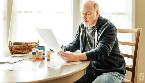 Man Sitting At Table Sorting Reading Paperwork with Prescription Medications