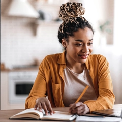 A woman sitting at a desk looking at her laptop and taking notes on paper.