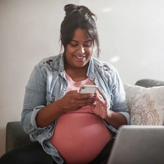 Pregnant member sitting on her couch while looking up information on her cellphone, with her laptop opened infront of her.