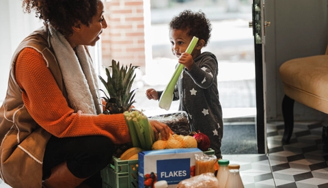Mother carrying groceries into her home while her young son holds onto a stalk of celery.