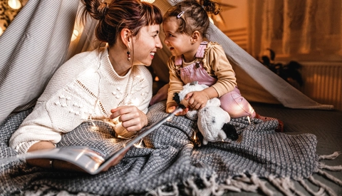 Mother and young daughter smiling and reading together in a makeshift fort