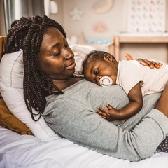 Mother relaxing in bed while her infant sleeps on her chest.