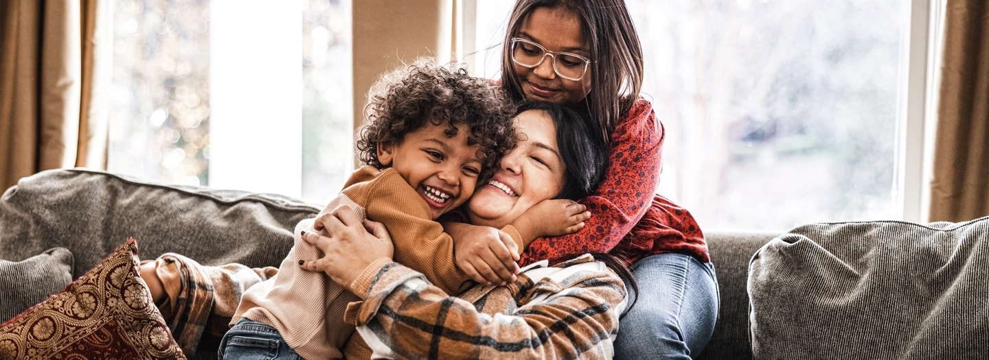 A mother sits on the couch hugging her two children
