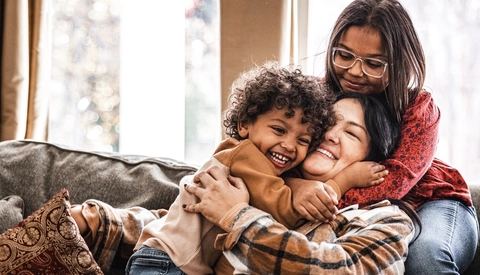 A mother sits on the couch hugging her two children