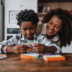 A child sits at a table playing with a push pop fidget toy while his caregiver is standing behind him smiling