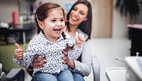 Toddler giving a thumbs up while sitting on her mother's lap at doctor's office