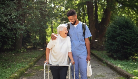Senior woman and caregiver outdoors on a walk with walker in park, talking and laughing