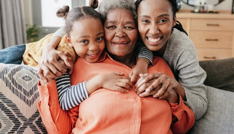 Cheerful granddaughter and mother hugging senior woman from behind and smiling at camera on sofa at home