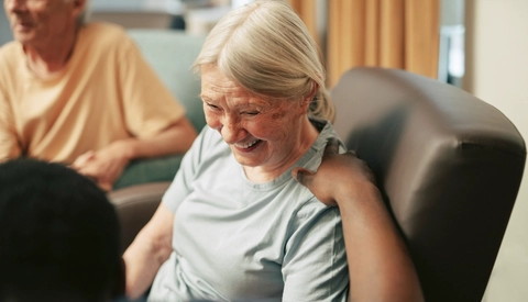Caregiver with hand on shoulder of happy senior woman sitting on armchair at retirement home