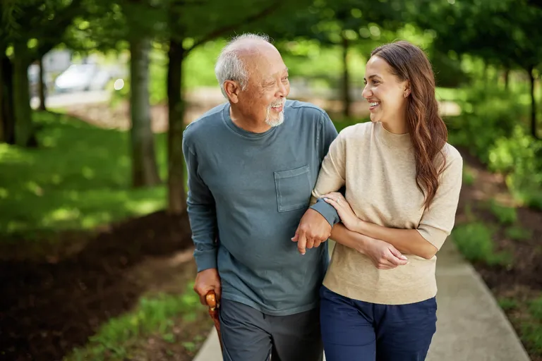 An older adult and a younger adult walk arm in arm along a tree-lined path outdoors. The pair are engaged with one another while walking, with lush green foliage and a softly blurred natural background creating a warm, inviting setting that suggests support, companionship, and active living.