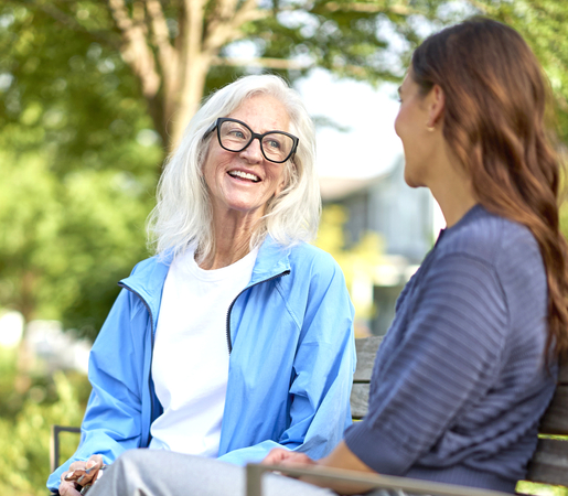 Senior and caregiver sitting on a bench and talking along a tree-lined walkway.