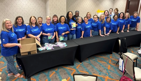 Volunteers in matching blue shirts stand behind tables assembling care kits during an indoor community service event.