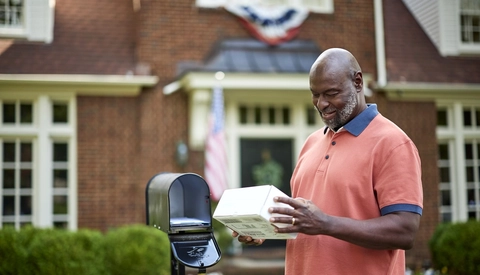 A person stands beside an open residential mailbox while holding a delivered package in front of a brick home