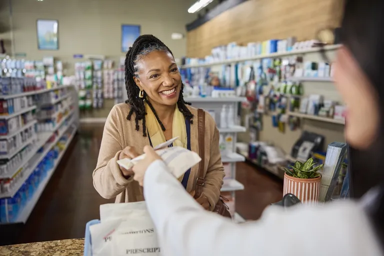 A pharmacy customer receives a prescription package from a pharmacy team member across a retail pharmacy counter.