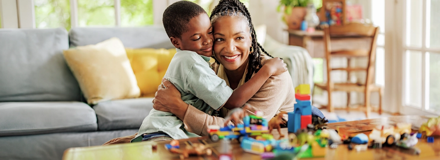 A senior adult and a child sit together on the floor of a bright living room, embracing near a coffee table covered with toys and building blocks. Large windows provide natural light, and the home setting emphasizes family connection, caregiving, quality time, and everyday life.