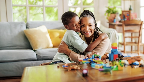 A senior adult and a child sit together on the floor of a bright living room, embracing near a coffee table covered with toys and building blocks. Large windows provide natural light, and the home setting emphasizes family connection, caregiving, quality time, and everyday life.