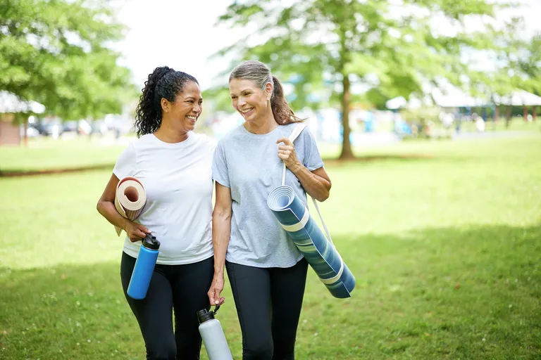 Two older adults walk together through a park carrying exercise mats and water bottles.