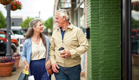 Older couple walking along a downtown sidewalk near local shops and storefronts. One person carries shopping bags while the other carries a beverage, illustrating an active and independent lifestyle in a community setting.