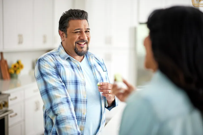 Older adult preparing a snack in a home kitchen while interacting with another adult. The image depicts a routine daily activity in a residential setting, highlighting nutrition, meal preparation, and independent living.