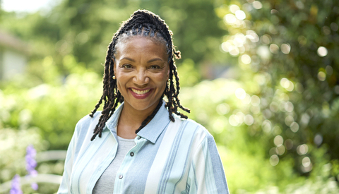 A person stands outdoors in a garden setting surrounded by greenery and flowering plants