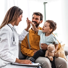 A caregiver sits on a couch with a provider, while holding a young boy in his lap. The child is holding a teddy bear and giving his medical provider a high five.
