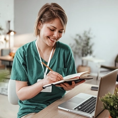 A medical professional sits in front of her laptop computer taking notes in a notebook.