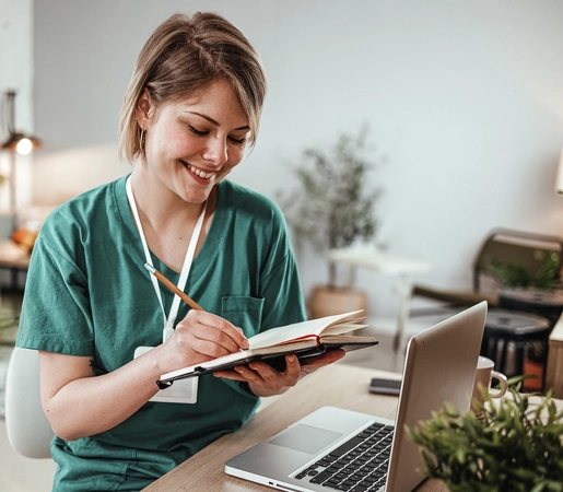 A medical professional sits in front of her laptop computer taking notes in a notebook.