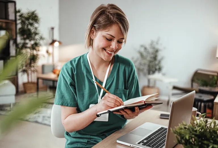 A medical professional sits in front of her laptop computer taking notes in a notebook.